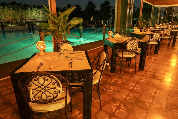 A restaurant with outdoor seating overlooking a pool, featuring tables with chairs and a potted plant.