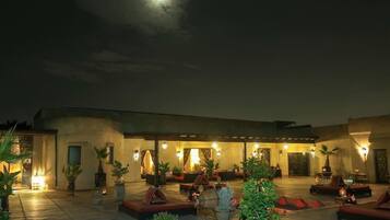 Un área de estar al aire libre con mesas y sillas, rodeada de plantas en macetas e iluminada por luces empotradas.
