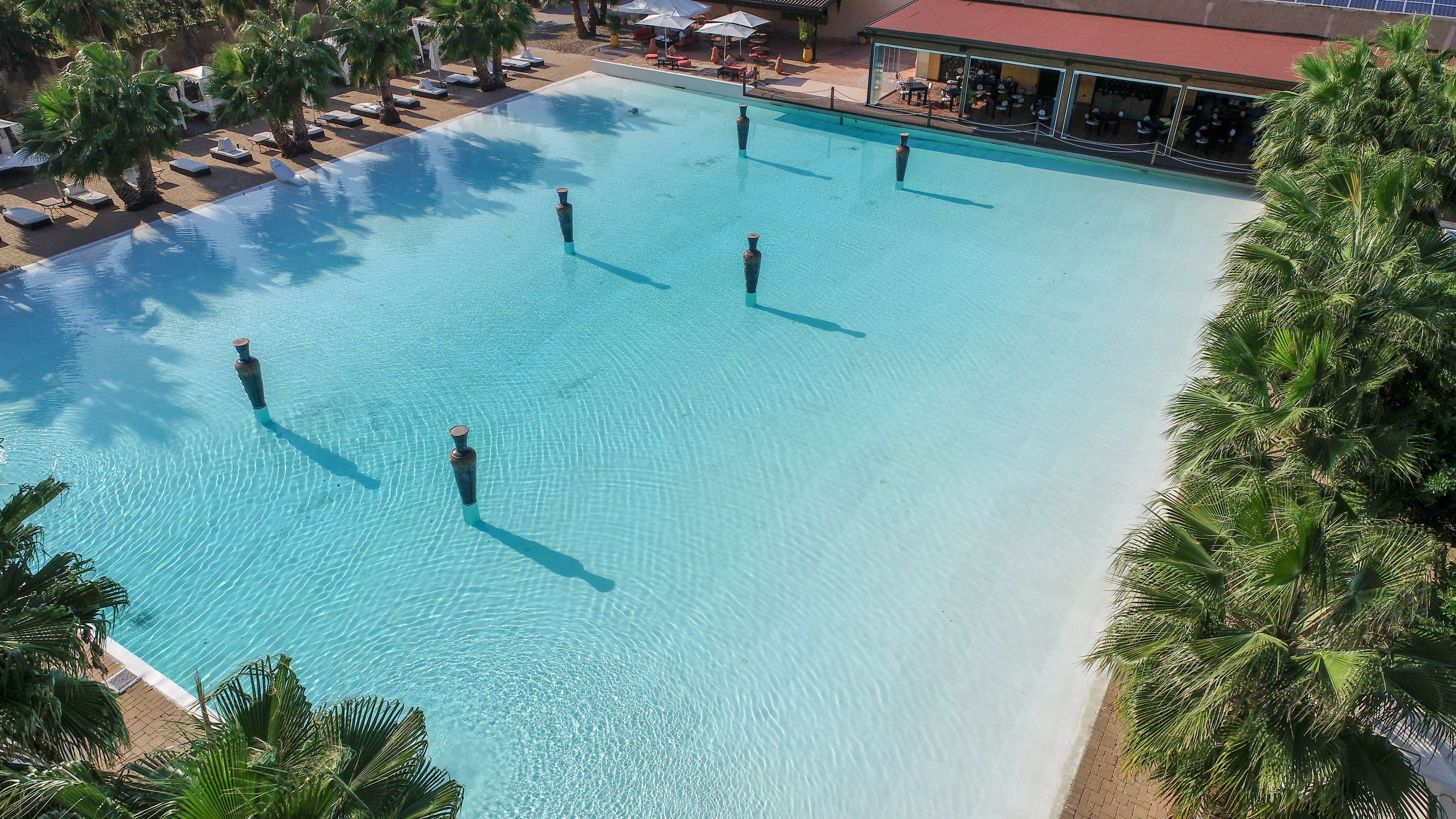 A large outdoor swimming pool with clear blue water, surrounded by palm trees and lounge chairs.