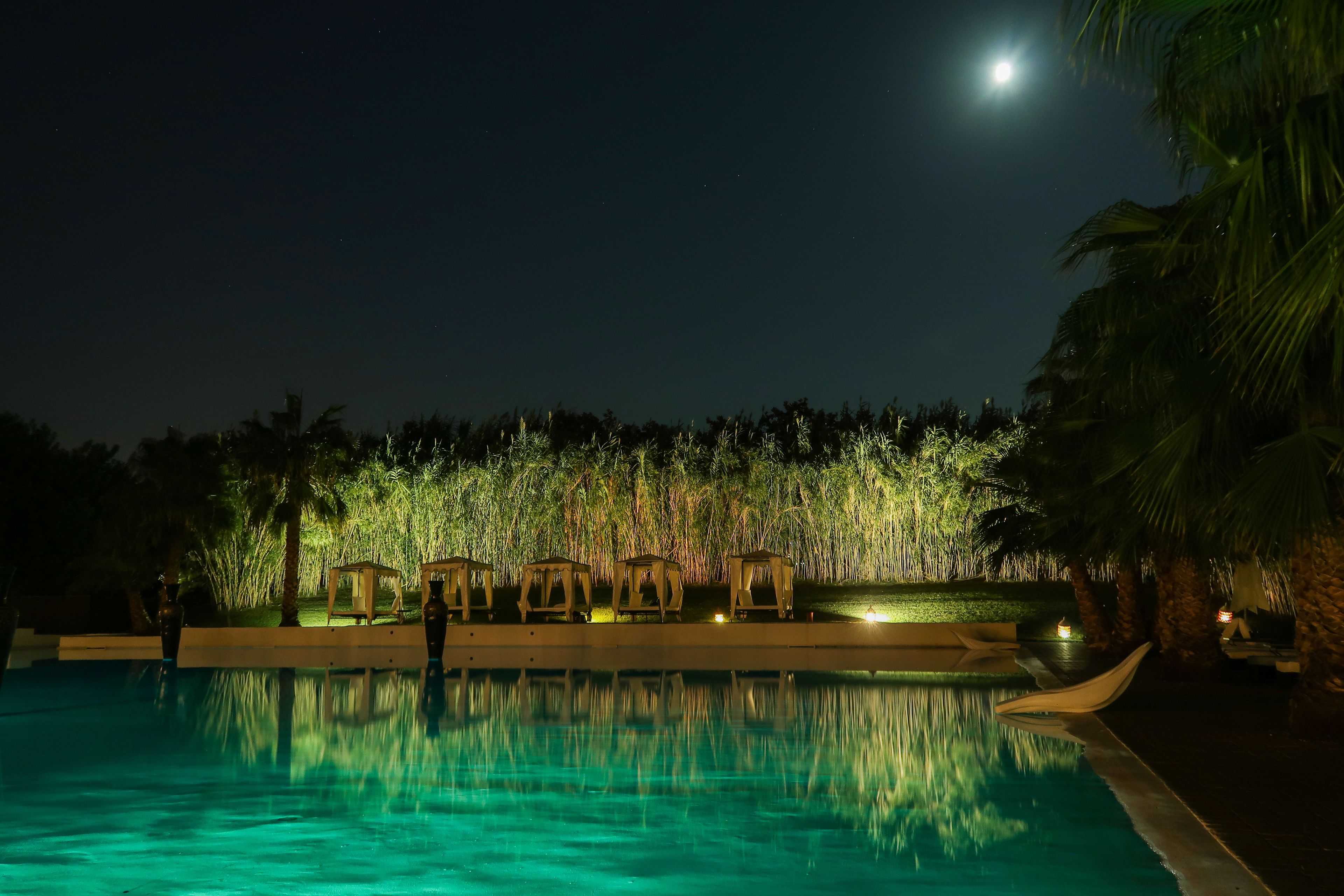 Un espace piscine nocturne offrant une vue dégagée sur la lune, une piscine paisible reflétant l’environnement, et une rangée de structures en bois le long du bord de la piscine.