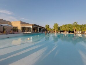 Una piscina con sillas de descanso y sombrillas, rodeada de palmeras y un edificio con grandes ventanales.