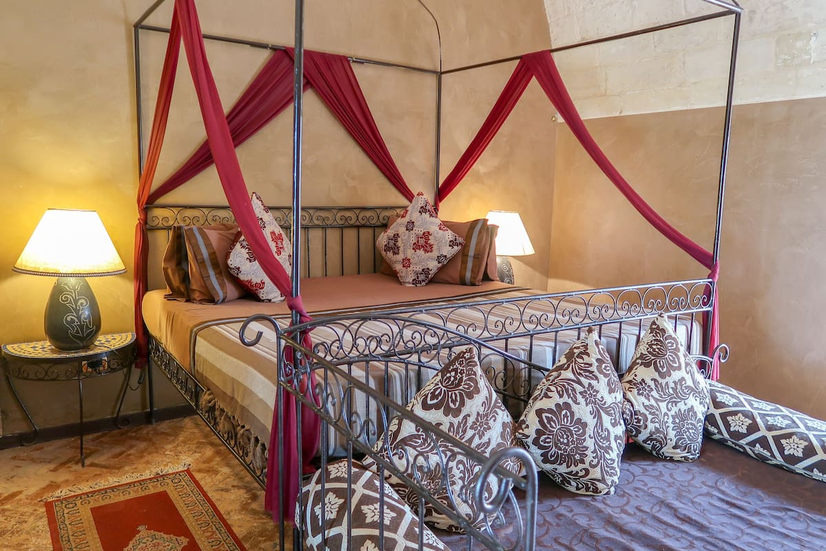 A four-poster bed with red drapes, patterned pillows, and two bedside lamps.
