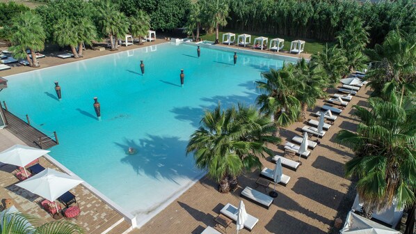 Una piscina rectangular grande con agua cristalina azul, rodeada de palmeras y sillas de descanso blancas.