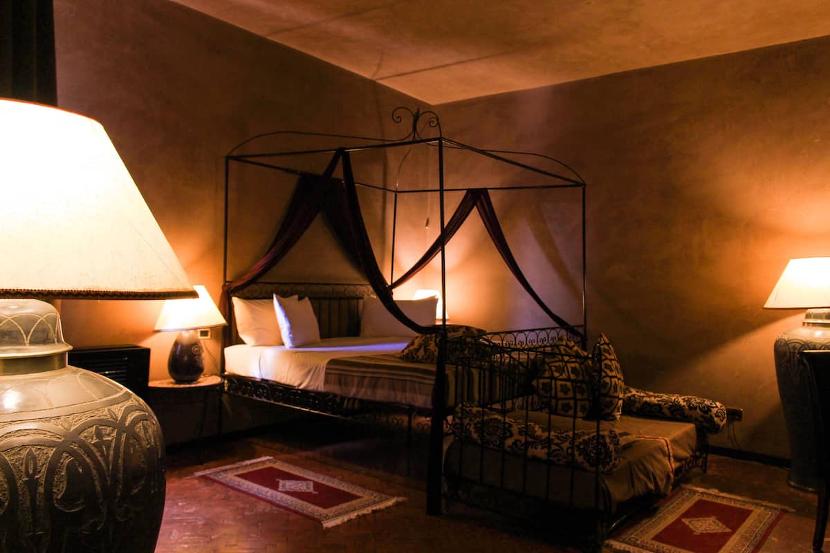 A dimly lit bedroom with a four-poster bed, two bedside lamps, a decorative vase, and a patterned rug.