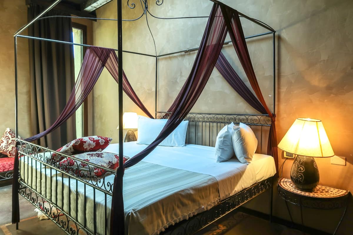 A four-poster bed with a canopy, a bedside table with a lamp, and a view of the outside through a window.