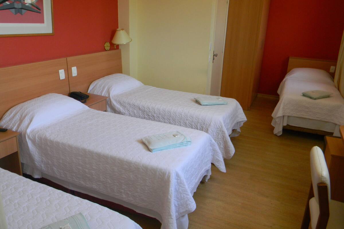 Ein Hotelzimmer mit drei Einzelbetten, einem Holzkopfstück, einem Nachttisch mit Lampe und einer Tür zu einem weiteren Raum.