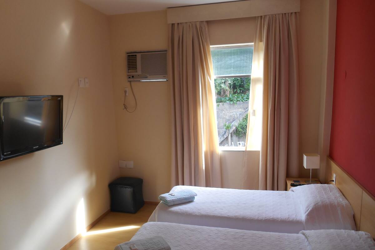 Ein Hotelzimmer mit zwei Betten, einem an der Wand montierten Fernseher, einer Klimaanlage, einem Fenster mit Vorhängen und einer kleinen Mülleimer.