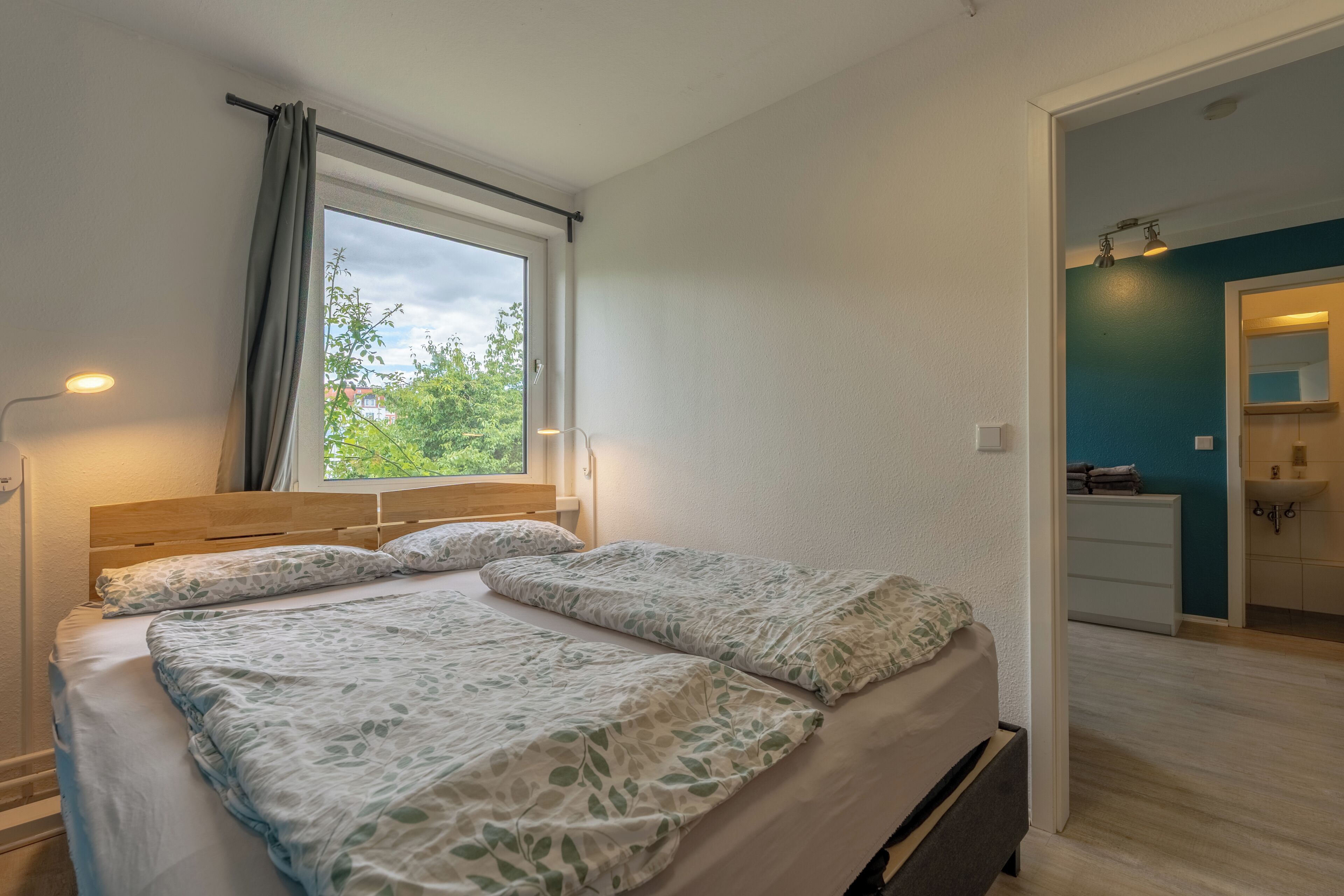 Un dormitorio con cama, ventana, lámpara de noche y vistas al exterior.