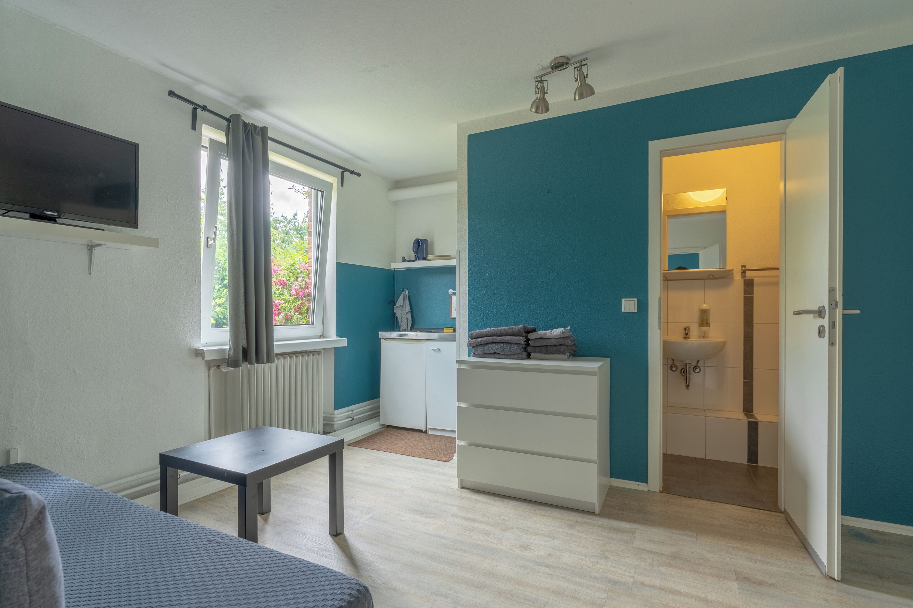 A compact room with a bed, a small table, a white wardrobe, a television, a window with curtains, a radiator, a sink, and a door leading to another room.