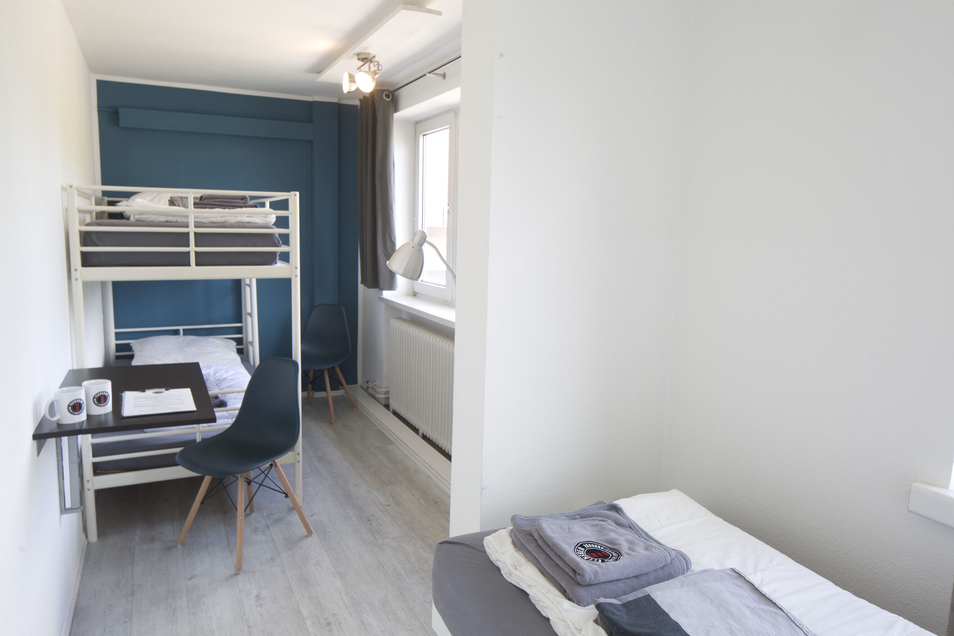 A dormitory room with bunk beds, a desk with a chair, a radiator, and a window with curtains.