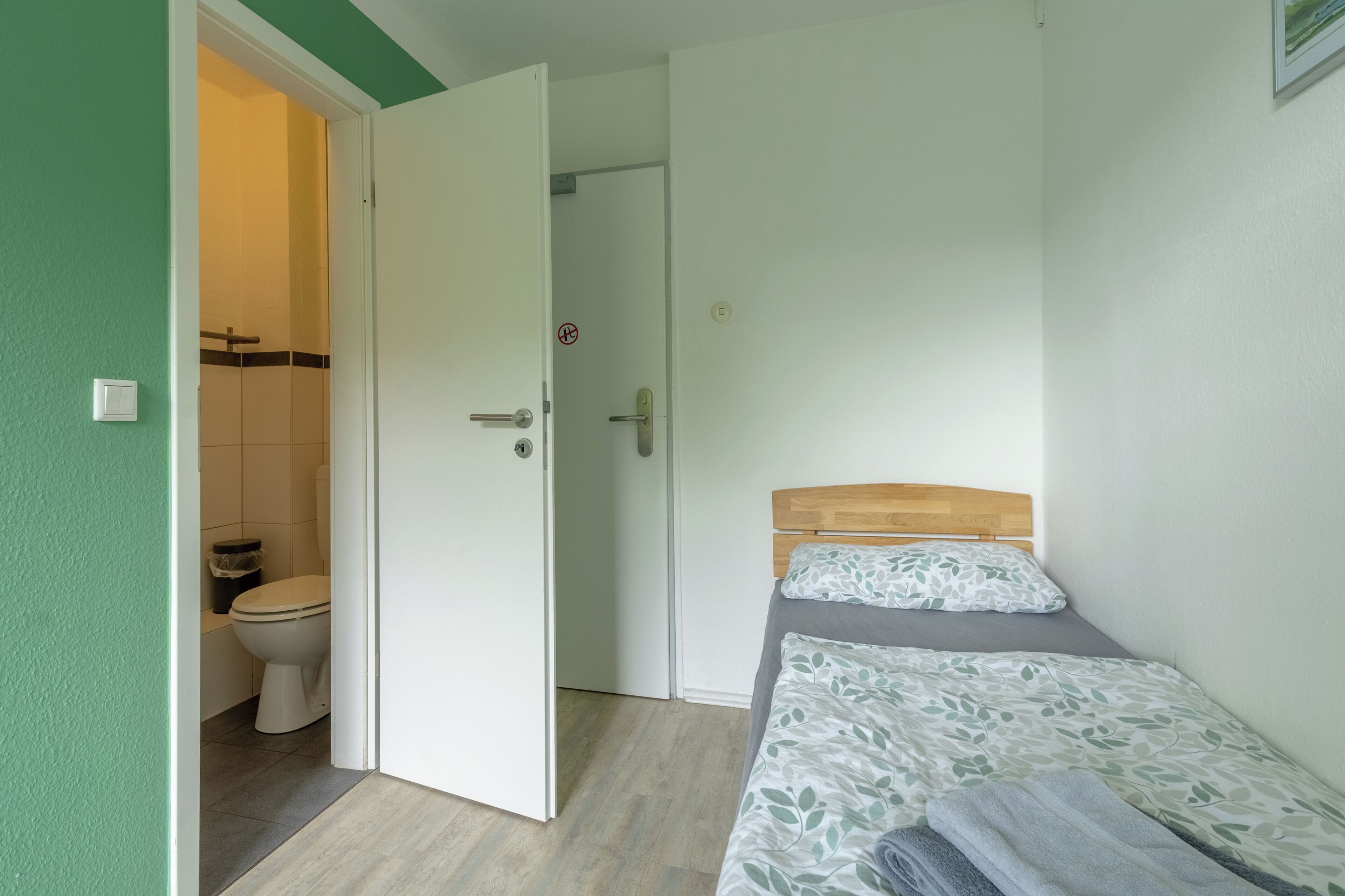 A small, single-room accommodation with a bed, a toilet, and a door leading to a bathroom.