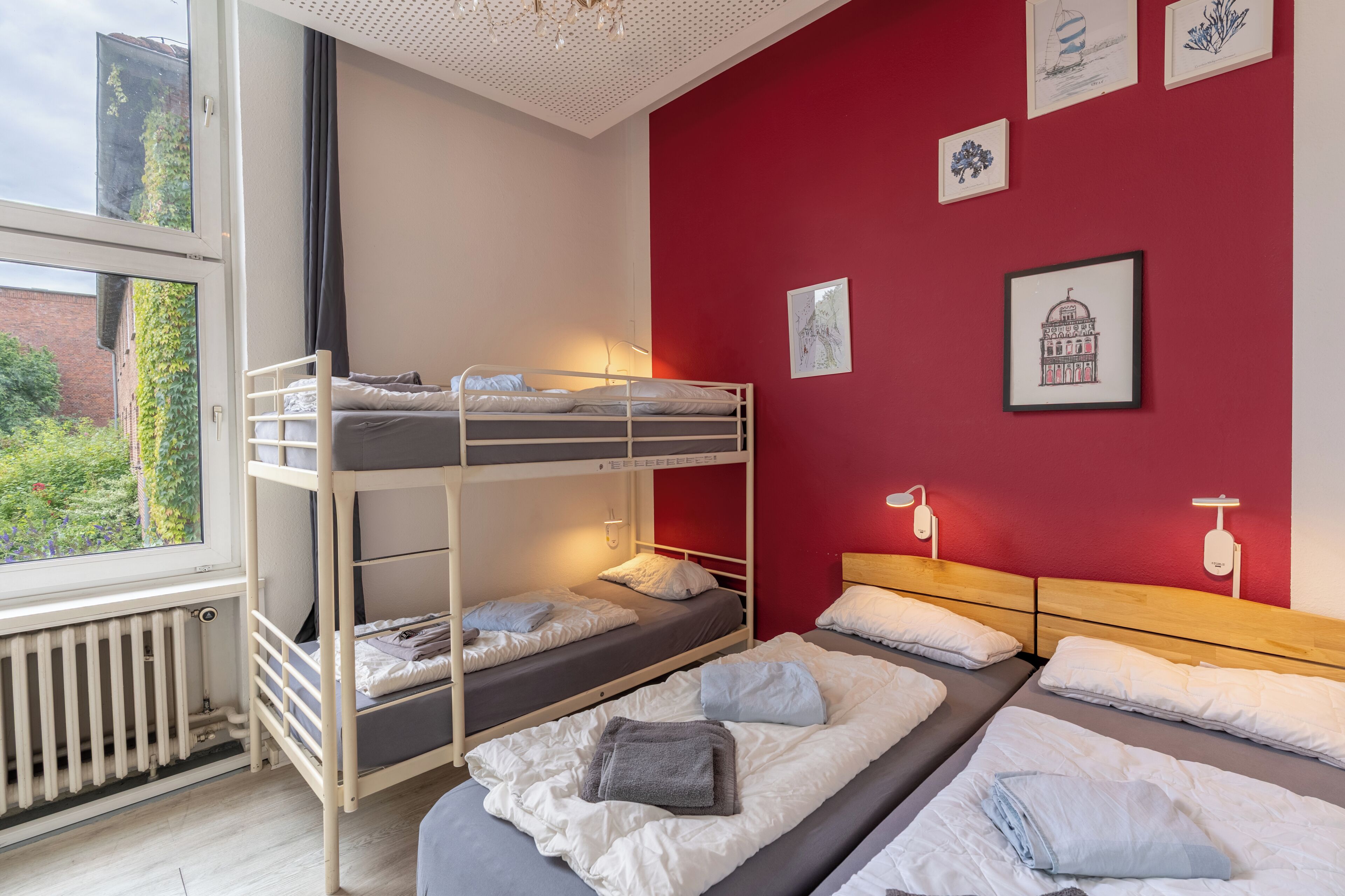 Een slaapzaal met stapelbedden, een radiator, een raam met uitzicht op groen en wanddecoratie.
