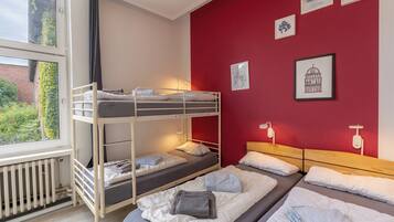 Een slaapzaal met stapelbedden, een radiator, een raam met uitzicht op groen en wanddecoratie.