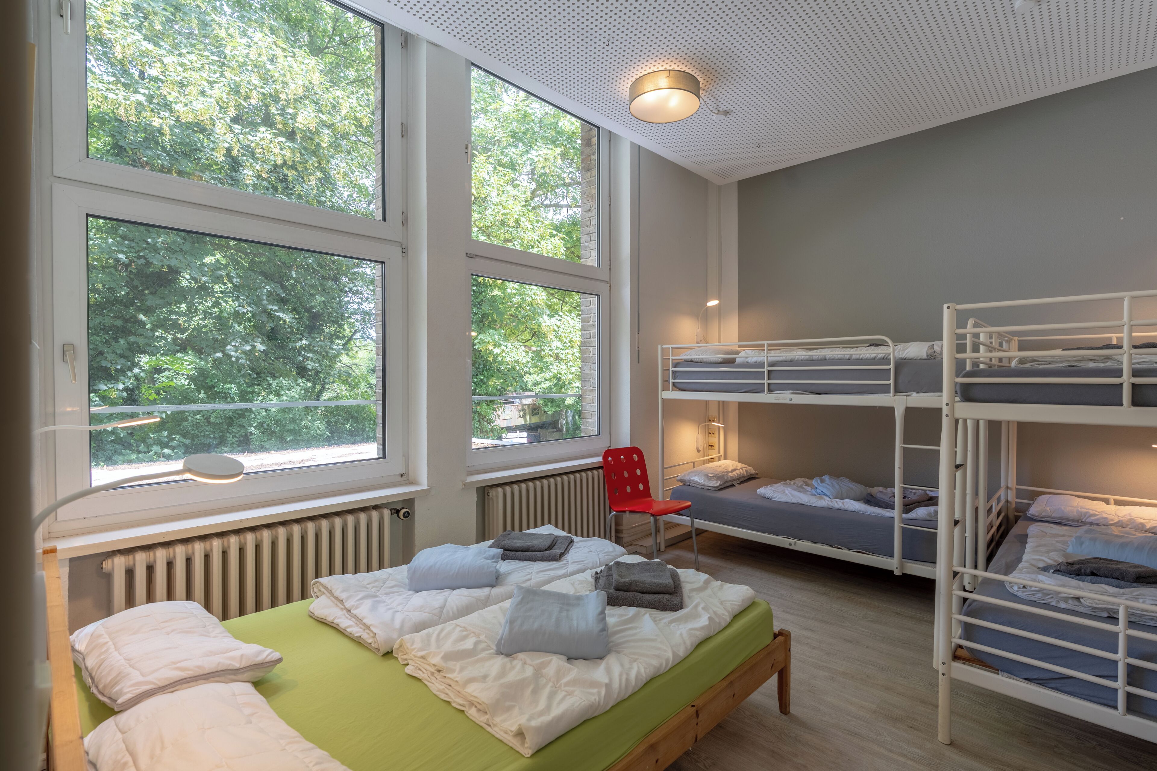 Habitación tipo dormitorio con literas, una cama individual, un radiador y ventanas amplias con vista a árboles.