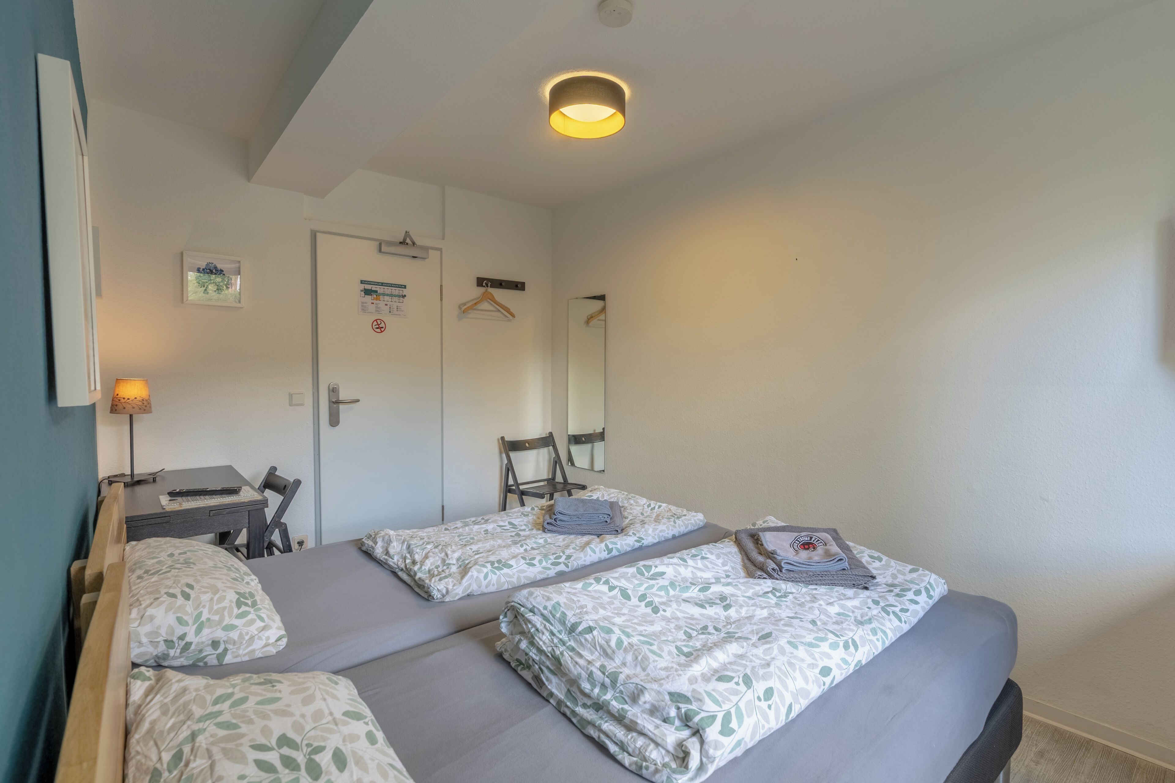 A room with two beds, a desk, a chair, a hanging coat rack, and a door with a notice.