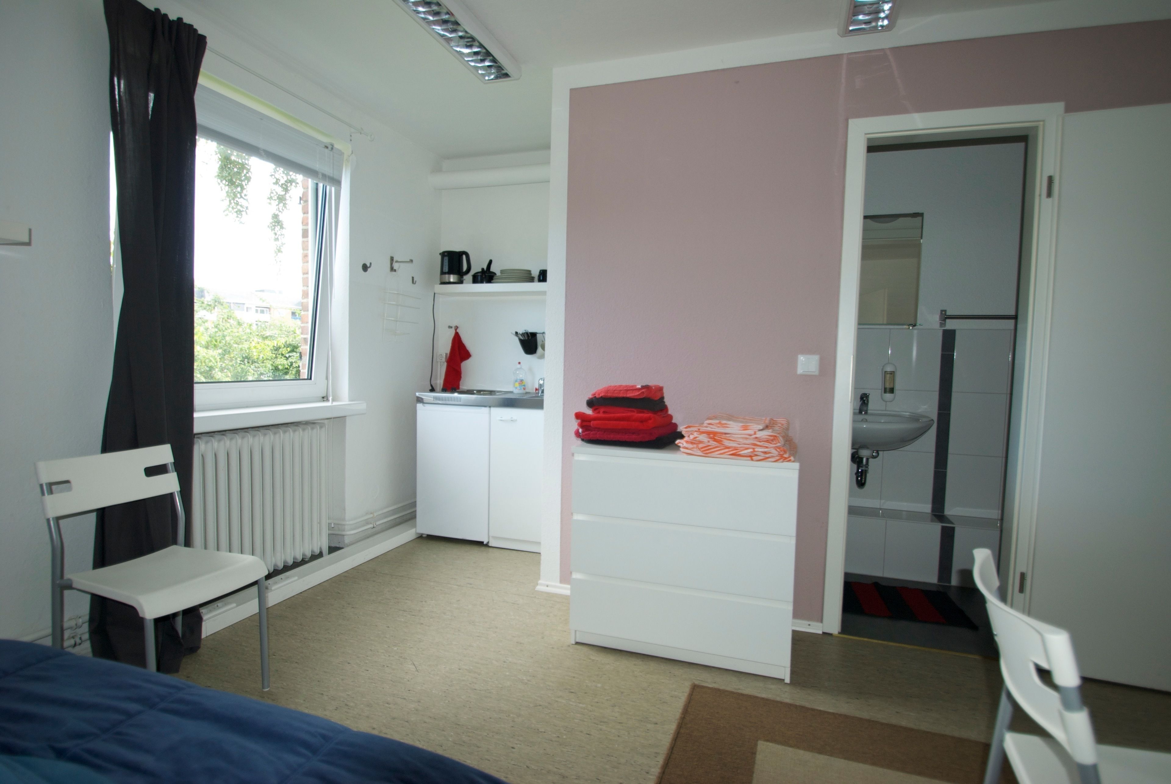 A room with a bed, a chair, a radiator, a white wardrobe, a sink, and a window with curtains.