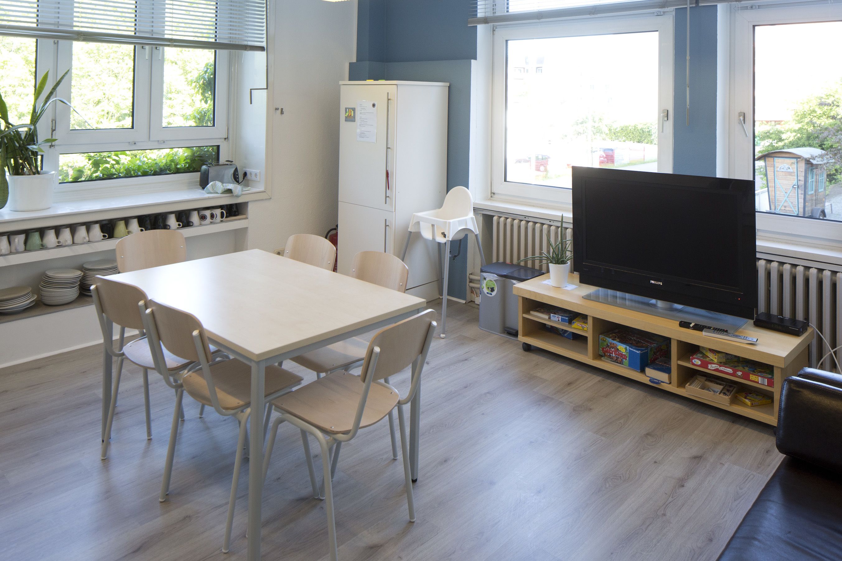 Eine moderne Küche mit Esstisch, Stühlen, einem Kühlschrank, einem Fernseher und einem Fenster mit Jalousien.