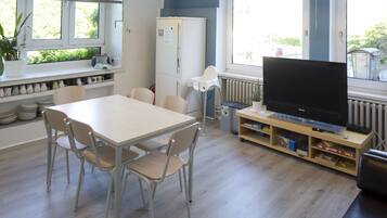 A modern kitchen with a dining table, chairs, a refrigerator, a television, and a window with blinds.