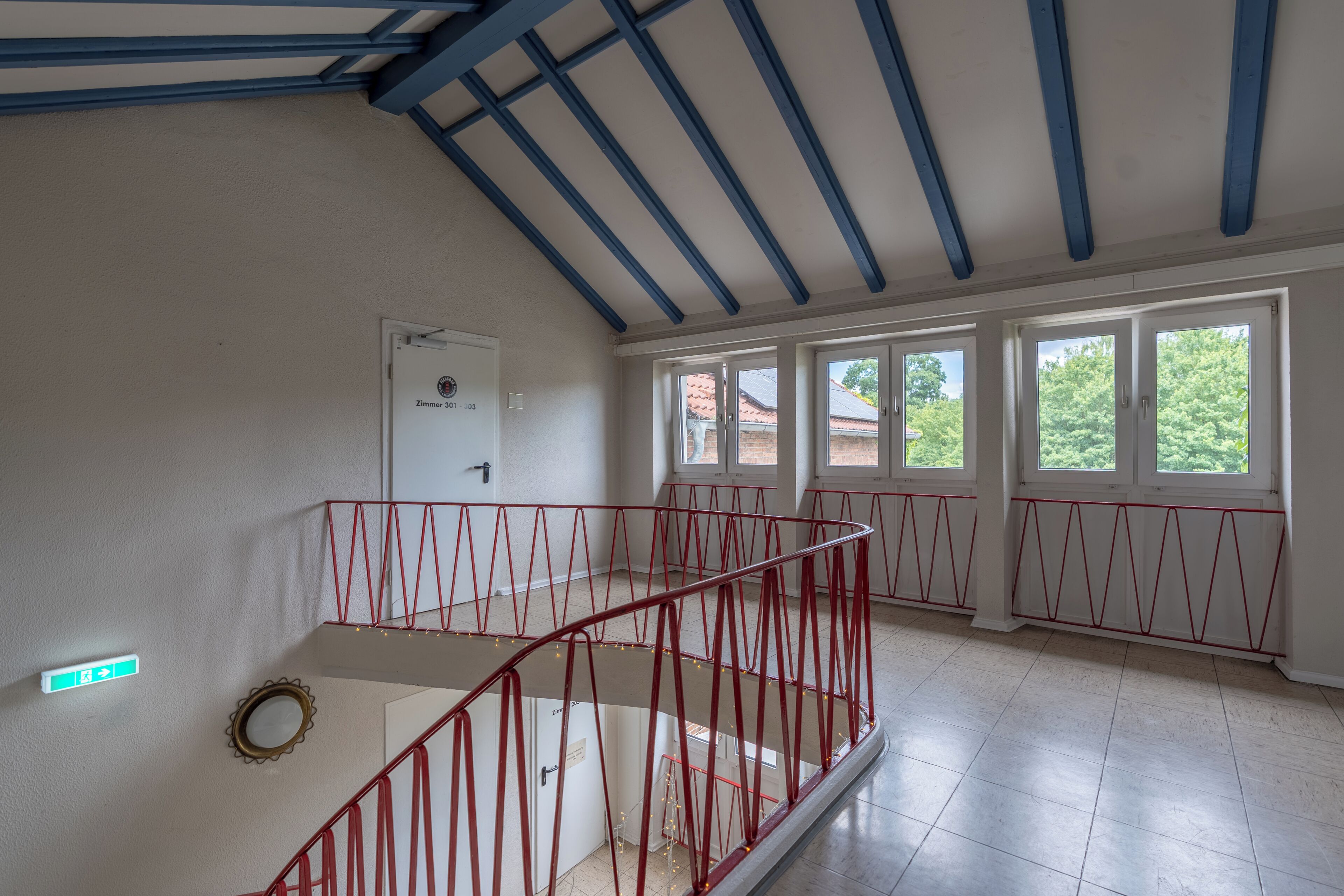 Una habitación amplia con techo alto, una escalera con barandillas rojas y grandes ventanales que ofrecen vistas al exterior.