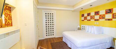 Habitación de hotel con cama, escritorio y silla, radiador, puerta con ventana y una pared decorada con obras de arte.