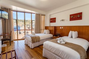 Habitación de hotel con dos camas, televisión, balcón con vistas a un paisaje urbano y suelo de madera.