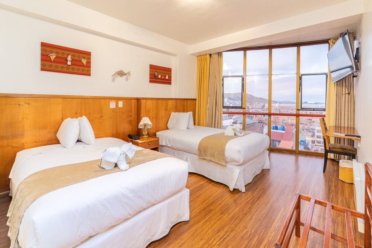 Ein Hotelzimmer mit zwei Betten, einem Holz-Kopfteil, einem Schreibtisch, einem Stuhl und einem großen Fenster mit Blick auf die Stadt.