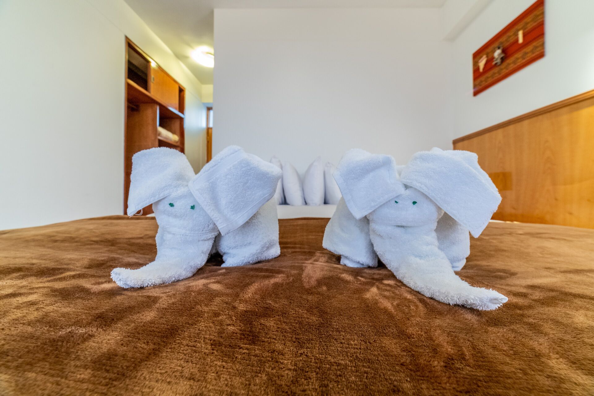 A hotel room with a brown carpet, a wooden headboard, and two white towels shaped like elephants.