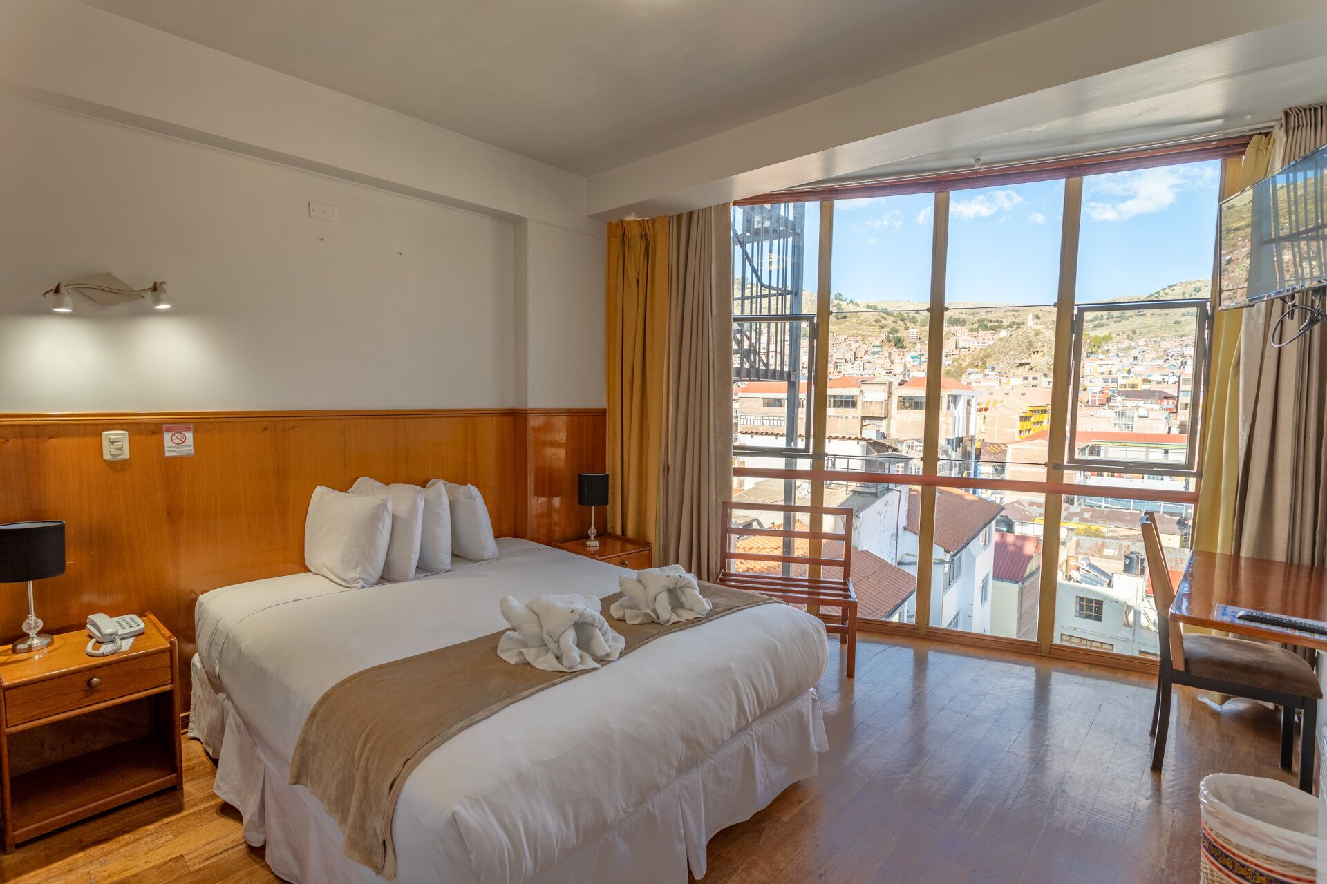 Habitació d'hotel amb un llit gran, una taula de nit de fusta amb un telèfon, un escriptori amb una cadira i una finestra gran que ofereix una vista de la ciutat.