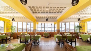 Un restaurante con varias mesas, manteles verdes, sillas de madera y vistas a la ciudad y colinas a través de amplias ventanas.