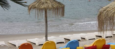 Una playa con sombrillas de paja y sillas de plástico de colores.