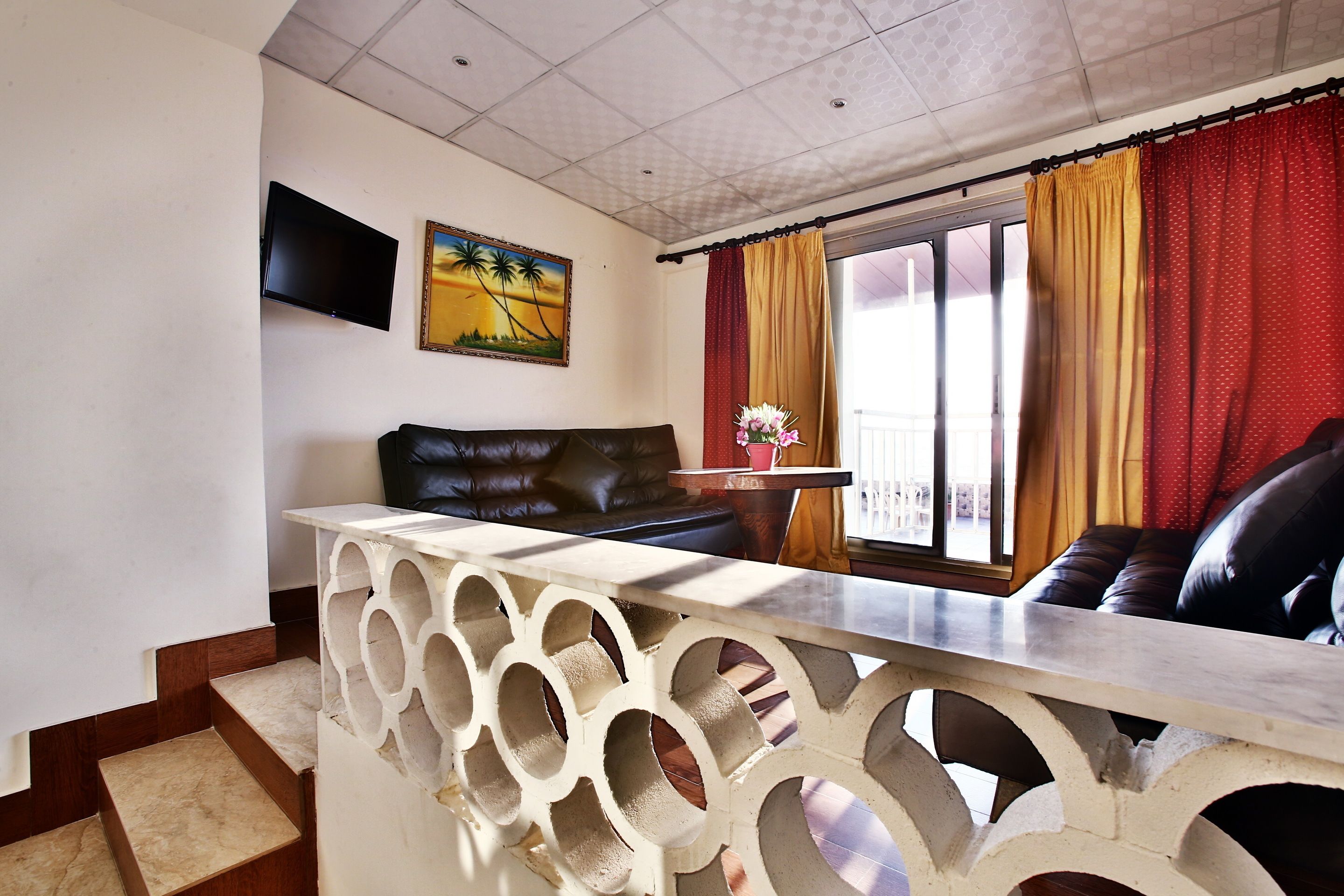 Un salon comprenant un canapé, un téléviseur fixé au mur, une fenêtre avec des rideaux et une petite table sur laquelle est posé un vase.
