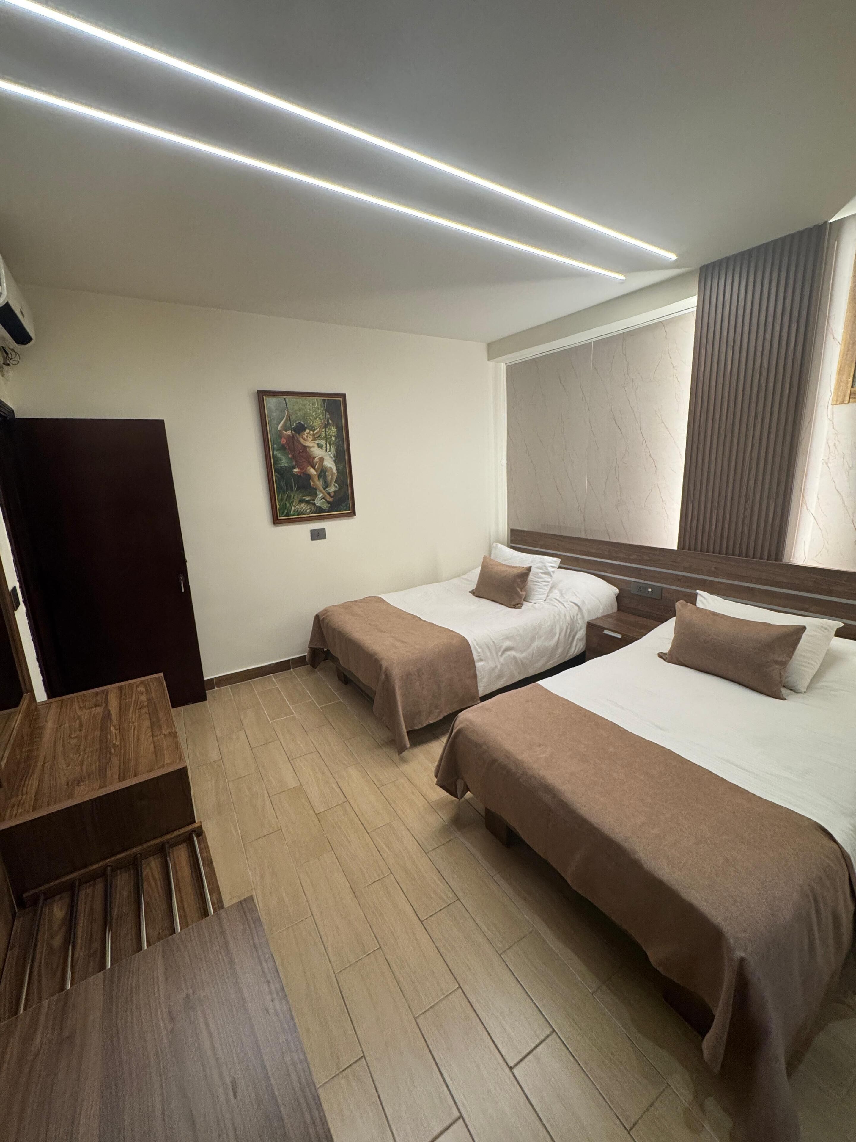 A hotel room with two beds, a wooden desk, a painting on the wall, and a large window with curtains.