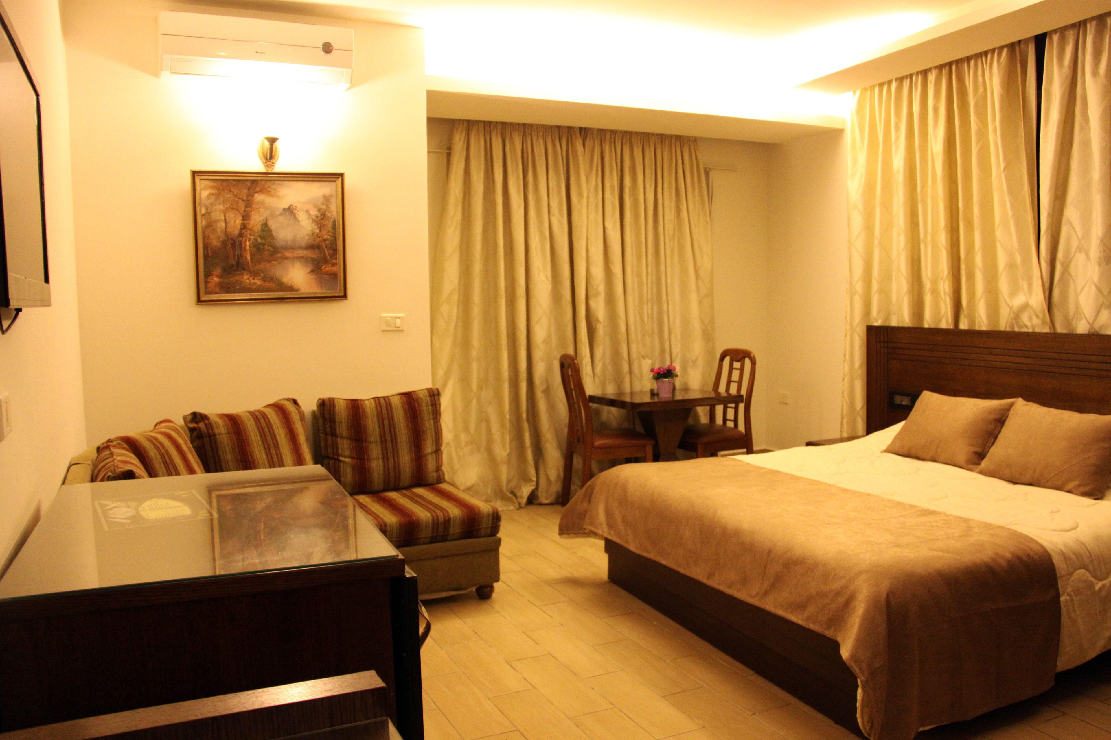 A hotel room with a bed, a sofa, a small table with a chair, a painting on the wall, and a window with curtains.