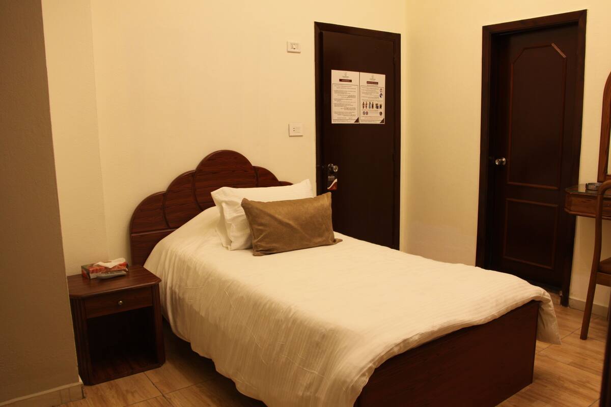Una cama bien hecha con cabecera de madera, una mesita de noche con un libro y una puerta con un aviso.