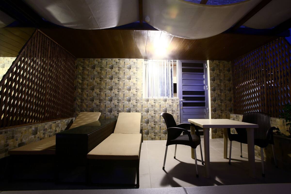 Una habitación con pared de piedra, un banco acolchado, una mesa con sillas y una ventana con cortinas.