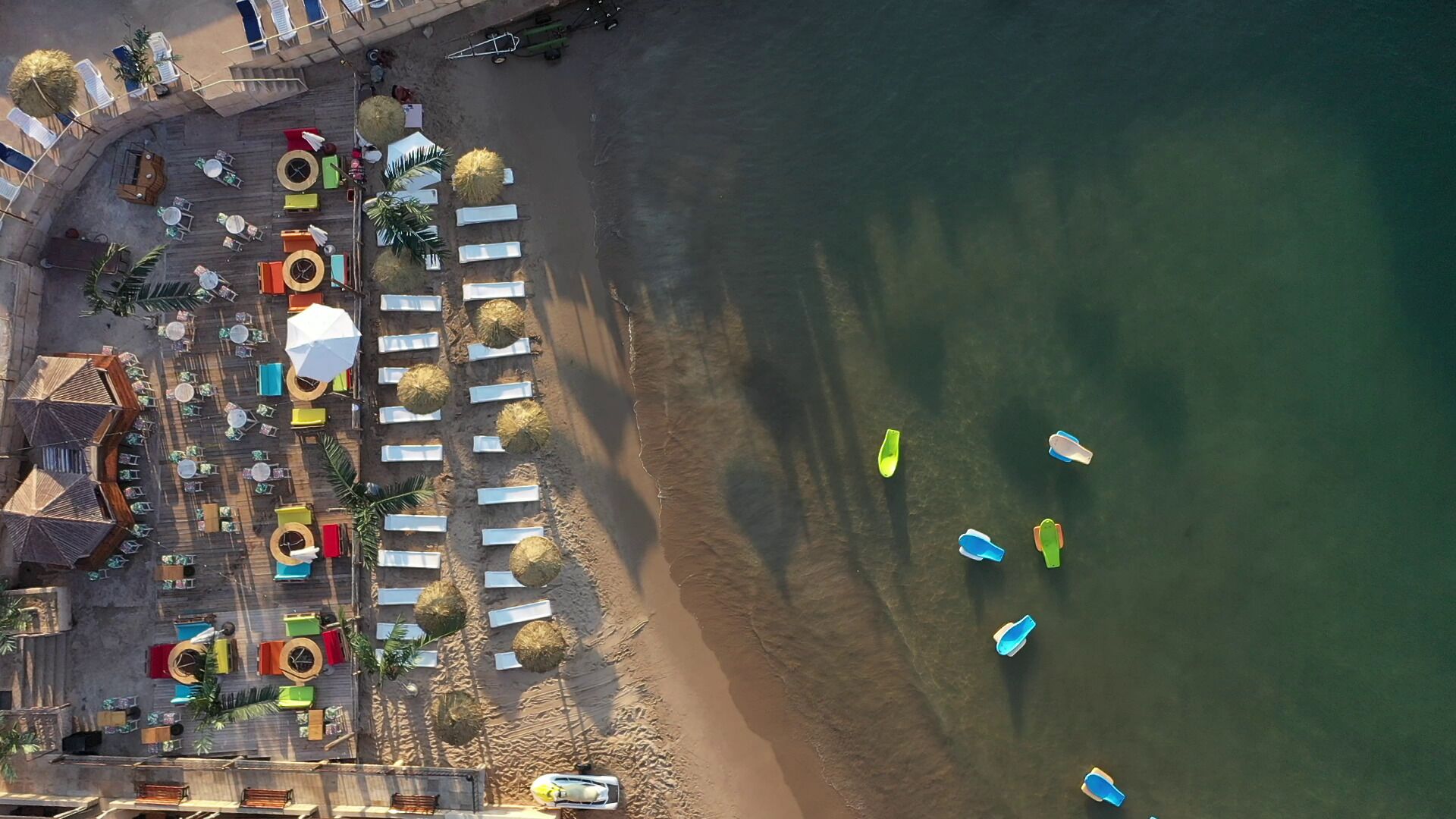 An aerial view of a beachside resort with rows of sun loungers and umbrellas, a sandy beach, and several boats in the water.