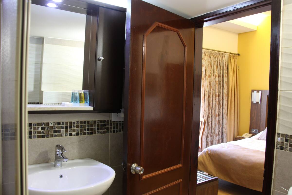 Baño con lavamanos, espejo y puerta de madera que da a un dormitorio con cama y cortinas.