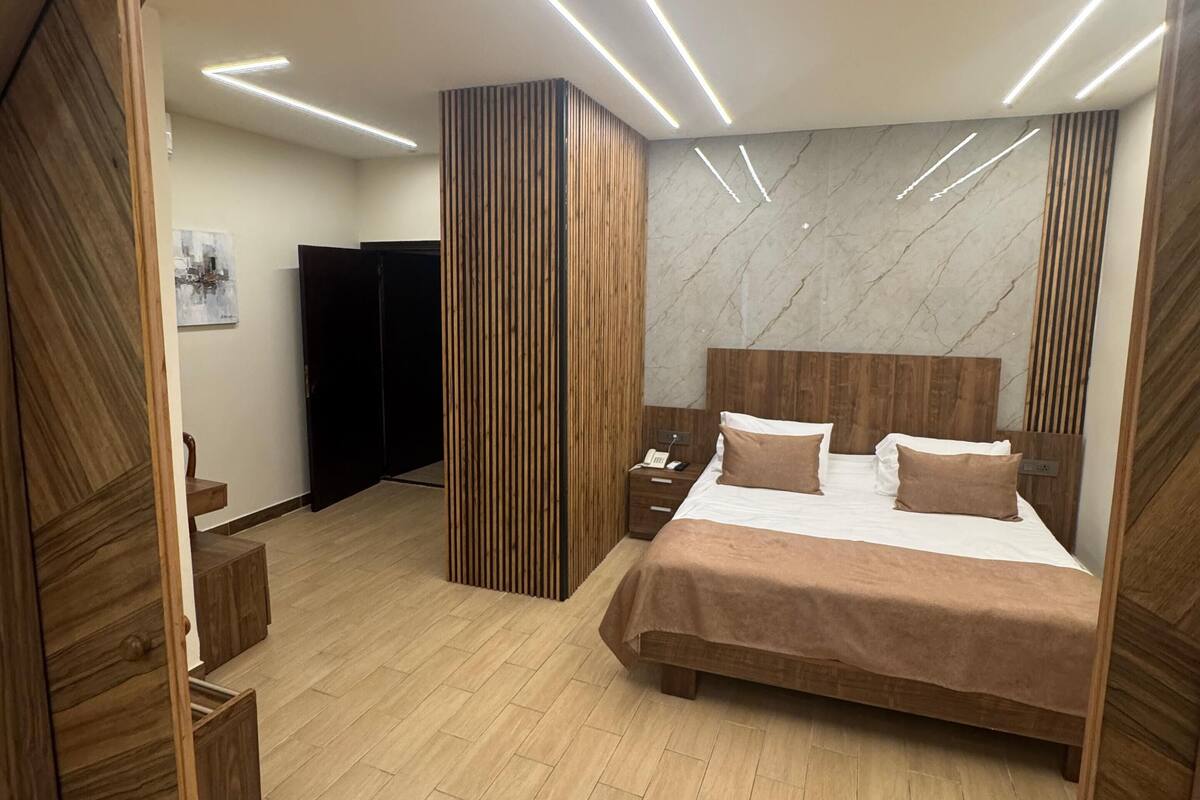 Un dormitorio moderno con cabecera de madera, una cama grande con muchas almohadas, una mesita de noche y un armario de madera.