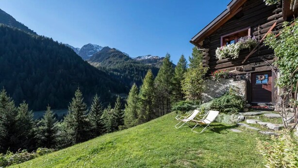 Een blokhut met uitzicht op bergen en een meer. Twee lounge fauteuils staan op het gras voor de hut.