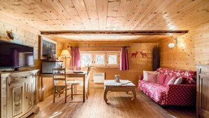 Een houten blokhut van binnen, met een televisie, een eettafel, stoelen, een bank en een raam met gordijnen.