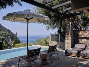 Zona junto a la piscina con vista al mar, sillas de madera, una tumbona y una pérgola con sombrilla.