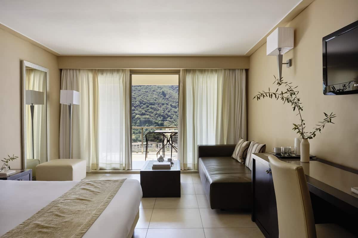 A hotel room with a bed, a sofa, a desk, a TV, and a view of a mountainous landscape.