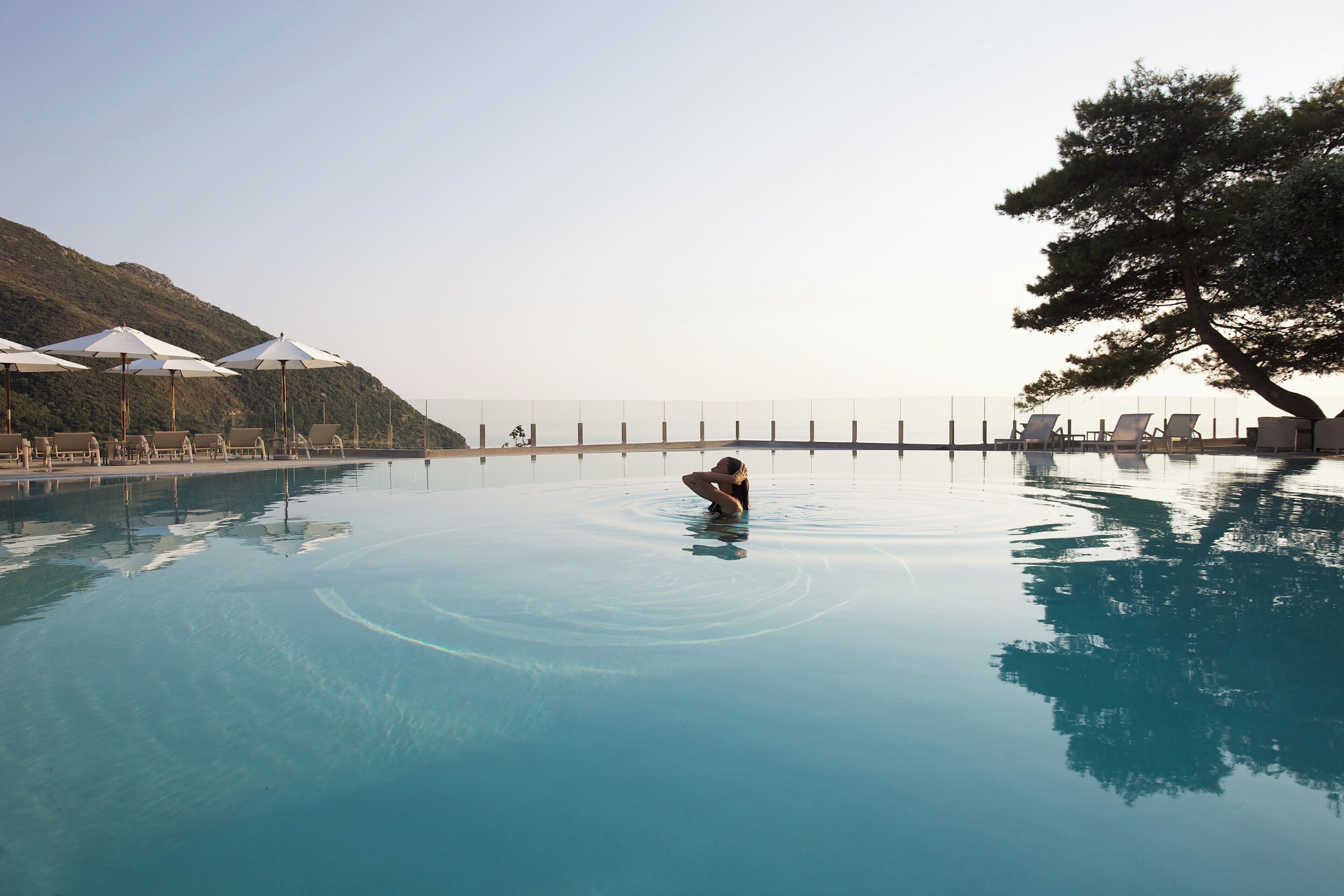 A swimming pool with lounge chairs and umbrellas, surrounded by a fence and a mountain backdrop.