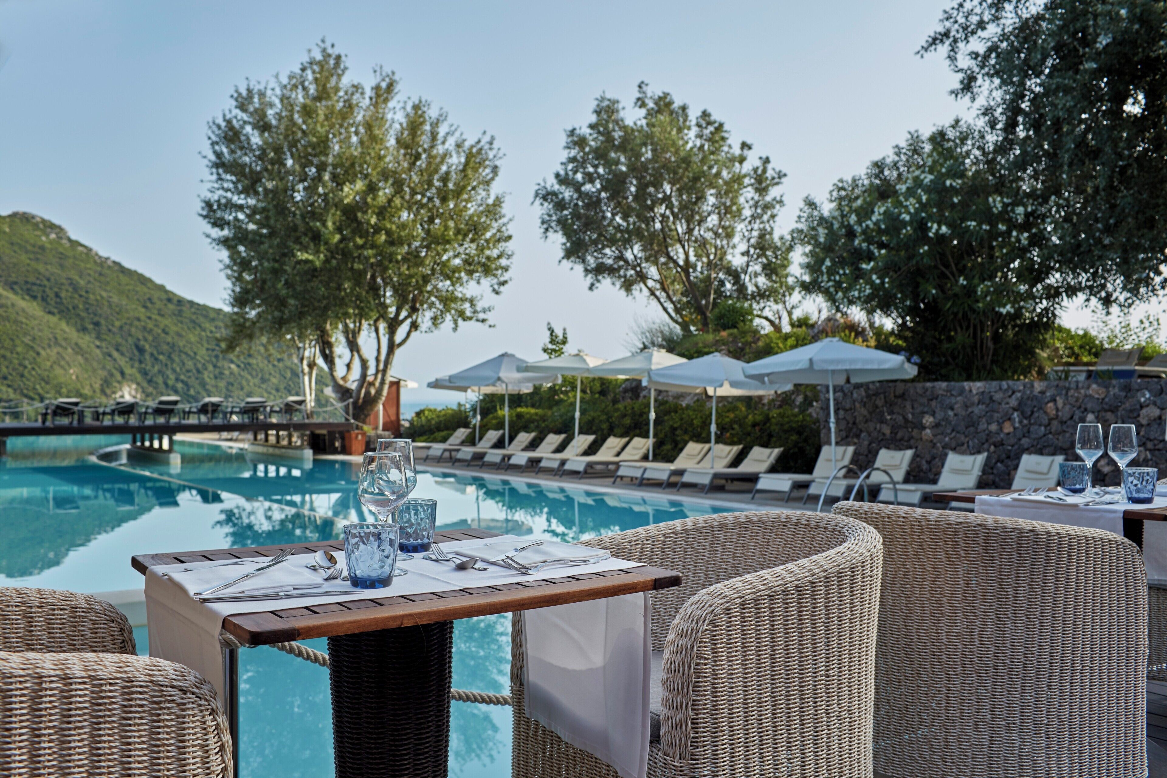 Zona de comedor junto a la piscina con mobiliario de mimbre, una mesa preparada para dos personas y sillones de descanso bajo sombrillas.