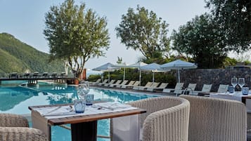 Zona de comedor junto a la piscina con mobiliario de mimbre, una mesa preparada para dos personas y sillones de descanso bajo sombrillas.