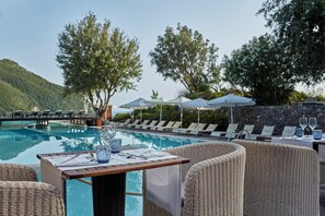 Zona de comedor junto a la piscina con mobiliario de mimbre, una mesa preparada para dos personas y sillones de descanso bajo sombrillas.