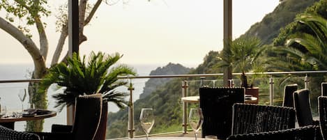 An outdoor dining area with wicker furniture, a table set for two, and a view of the ocean and hills.