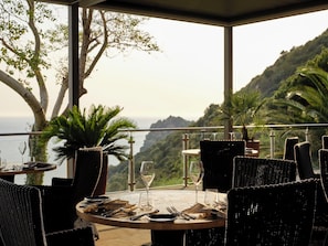 An outdoor dining area with wicker furniture, a table set for two, and a view of the ocean and hills.