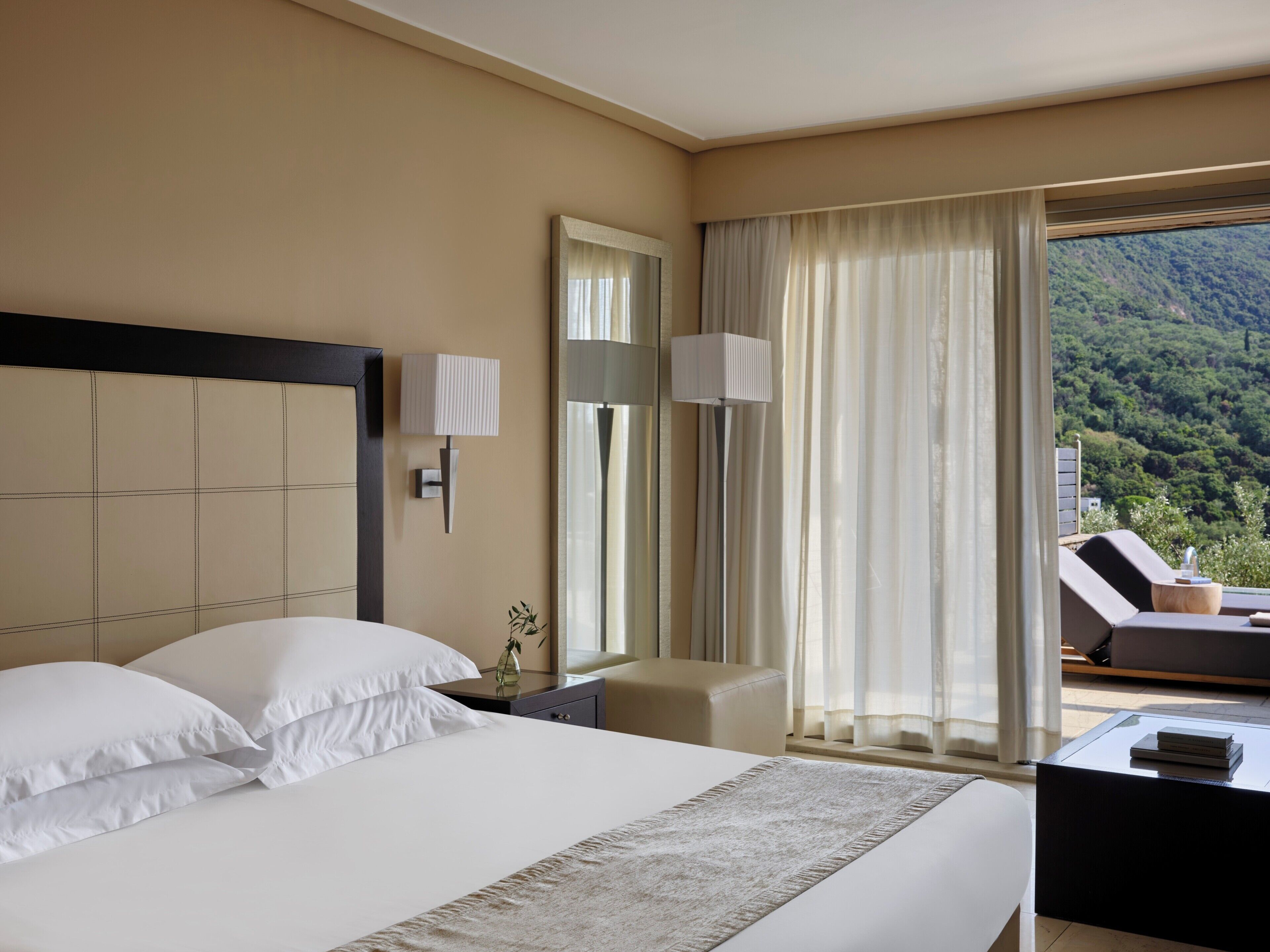 Een hotelkamer met een bed, nachtkastje, lamp, stoel en uitzicht op een bergachtig landschap.