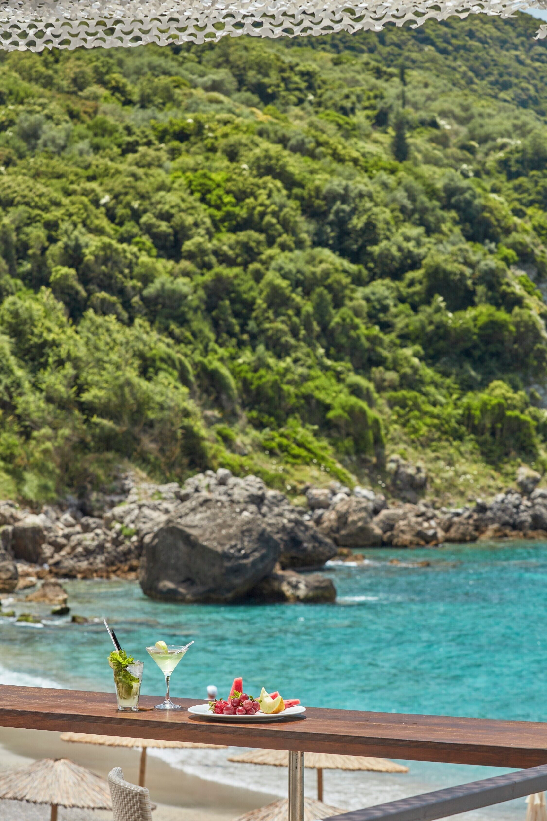A wooden table with a plate of fruit and two drinks, overlooking a turquoise sea and a forested hillside.